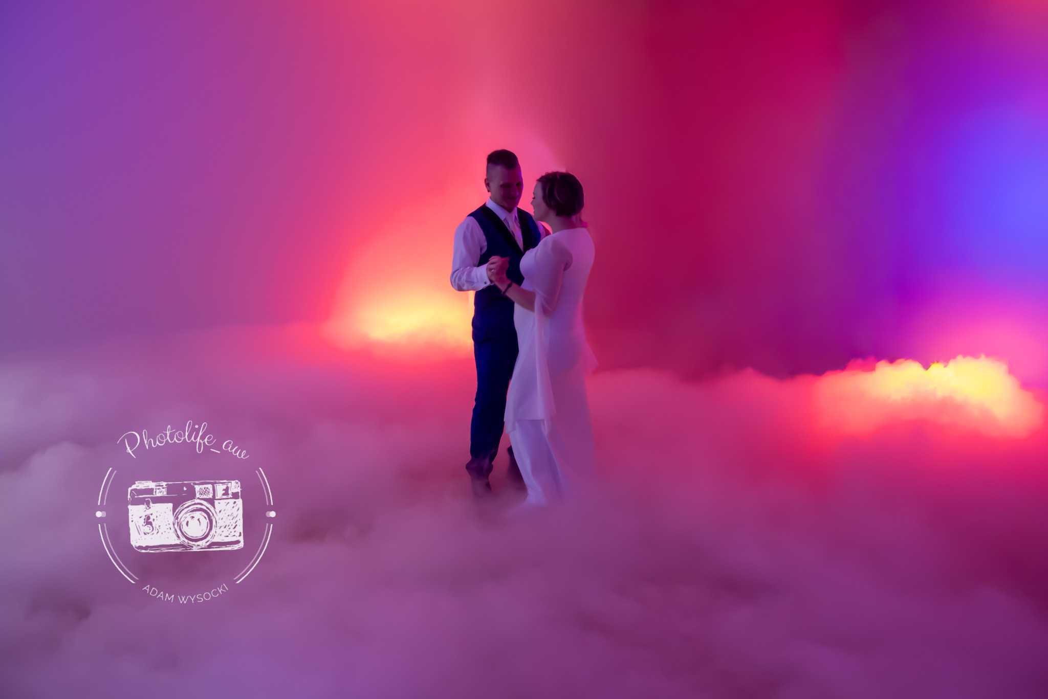 Para młoda w tańcu, otoczona sztuczną mgłą i kolorowym oświetleniem w odcieniach różu i fioletu. Widoczny logotyp fotografa w lewym dolnym rogu.
