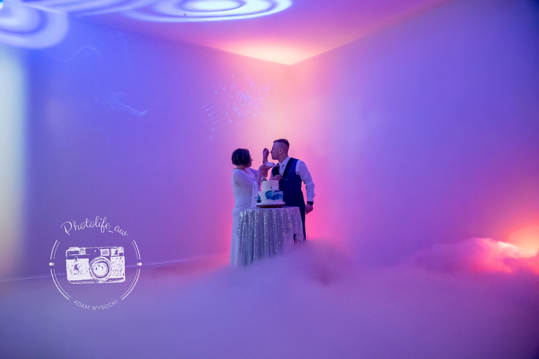 Para młoda kroi tort weselny w otoczeniu mgły i kolorowych świateł w Warszawie. Dekoracja stołu srebrną tkaniną. Ujęcie z logo fotografa.
