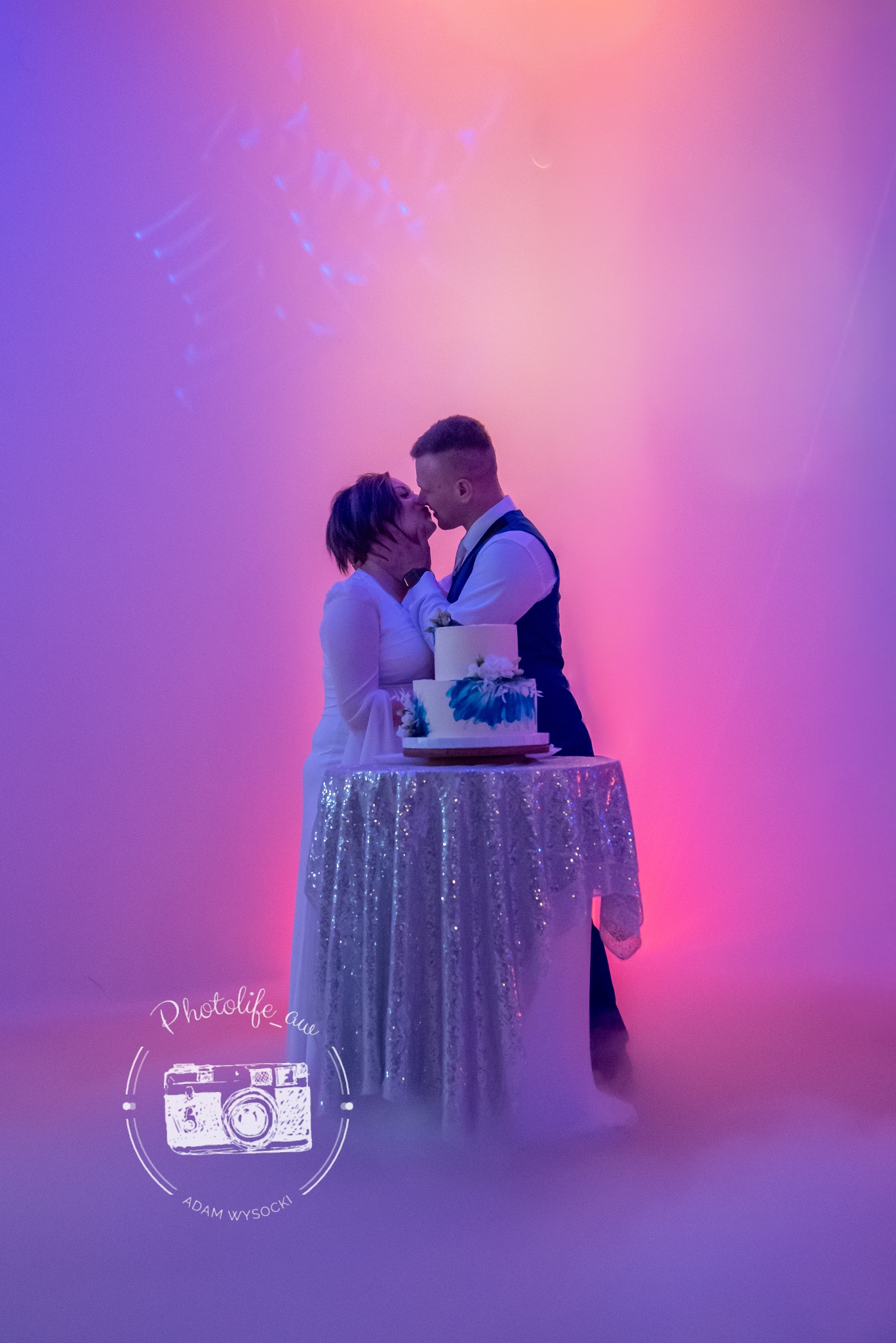 Pocałunek pary młodej przy torcie weselnym na tle fioletowo-różowego oświetlenia i mgły. Tort dwupiętrowy, dekorowany kwiatami. Stół przykryty błyszczącą tkaniną.