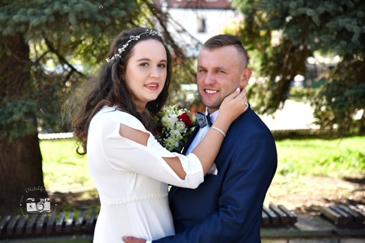 Uśmiechnięta para młoda w plenerze: panna młoda w białej sukni z kwiatami, pan młody w granatowym garniturze z muszką, delikatny uścisk dłoni na szyi.