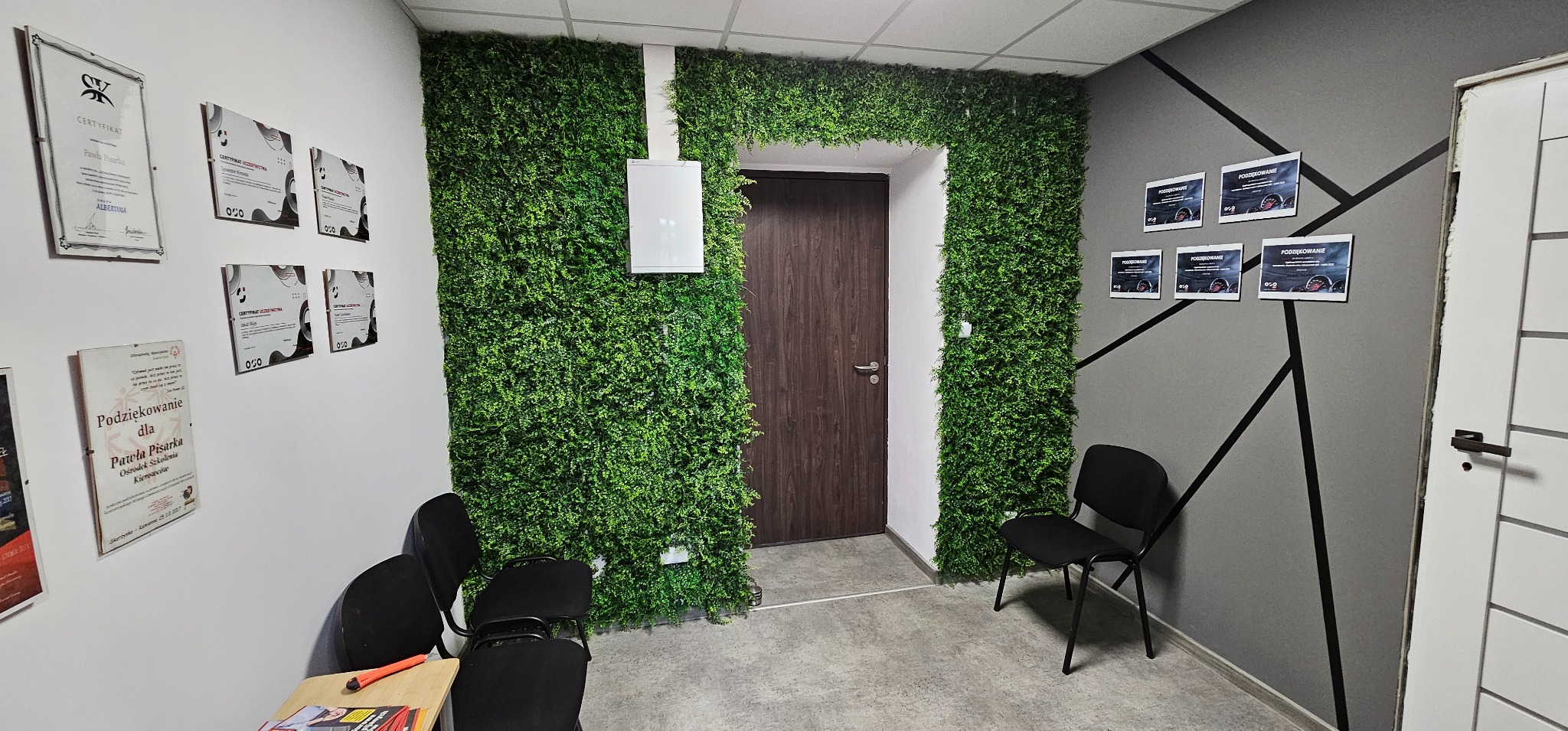 Wnętrze szkoły jazdy: ściana z certyfikatami, podziękowaniami, zielona ściana z roślin i drzwi. Krzesła dla kursantów w poczekalni.