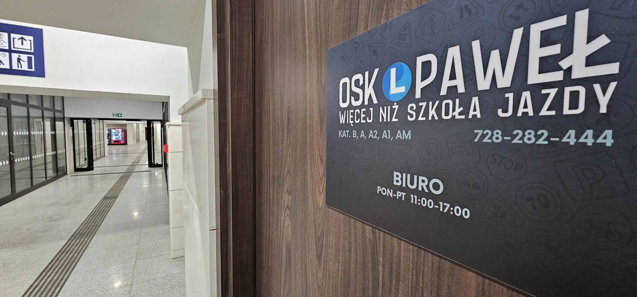 Tabliczka informacyjna szkoły jazdy OSK Paweł z numerem telefonu i godzinami otwarcia biura, umieszczona na drewnianej ścianie w jasnym korytarzu.