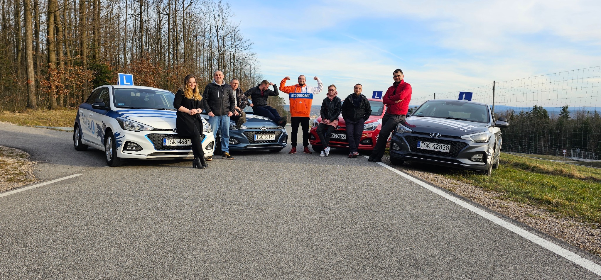 Grupa kursantów z instruktorami pozuje przy trzech samochodach oznaczonych symbolem 'L' na tle leśnego krajobrazu i błękitnego nieba. Jeden z kursantów prezentuje muskuły.