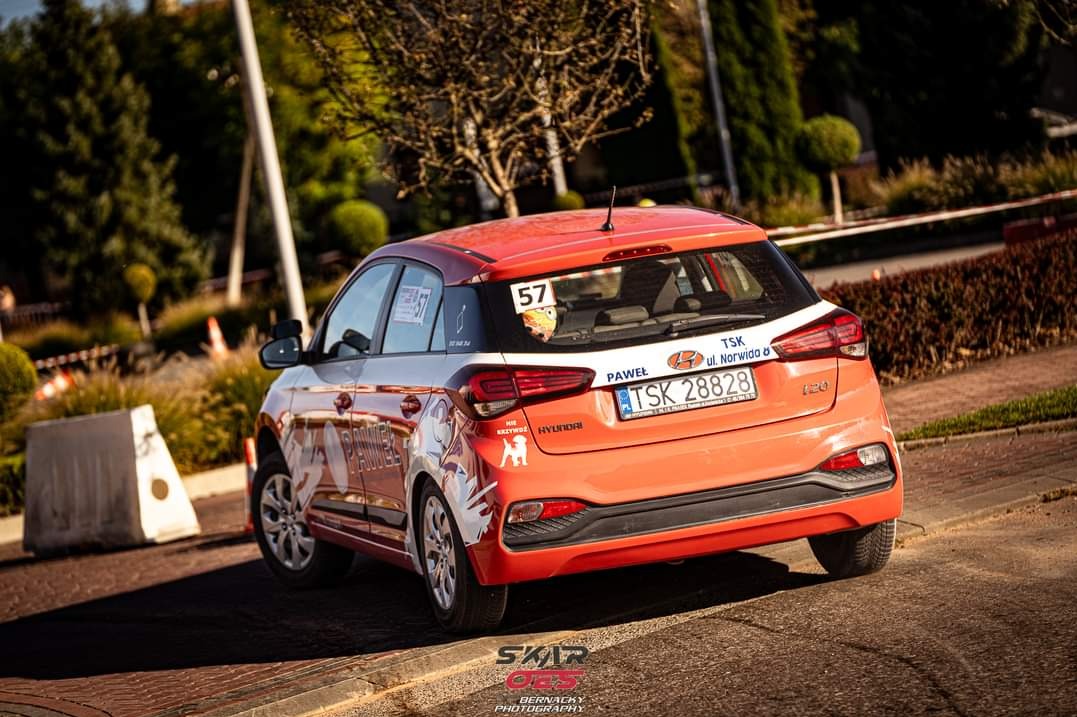 Pomarańczowy Hyundai i20 z numerem 57, oklejony grafikami, zaparkowany na ulicy. Widoczny tył pojazdu i otoczenie miejskie.