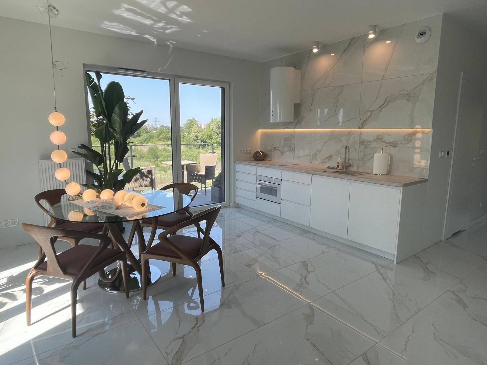 Jasna kuchnia z marmurową ścianą i białymi szafkami, oświetlona listwą LED. Na pierwszym planie szklany stół z drewnianymi krzesłami i dekoracyjną lampą.