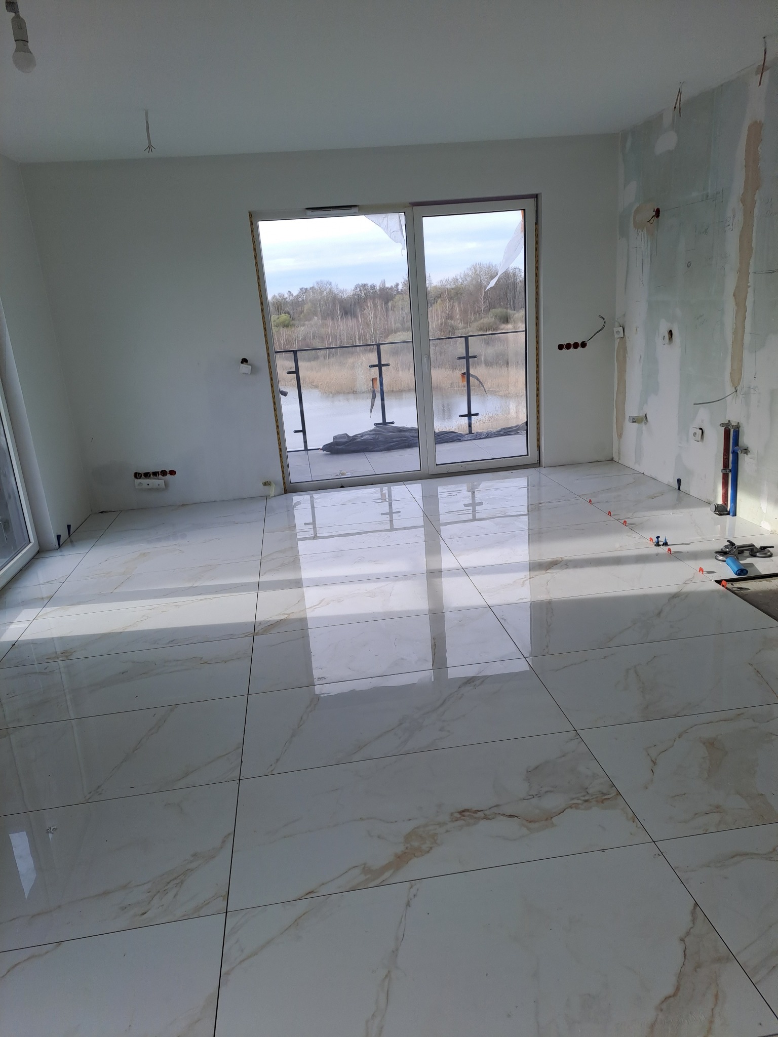 Połyskujące, marmurowe płytki podłogowe w jasnym pomieszczeniu z widokiem na jezioro przez duże okno balkonowe. Częściowo wykończona ściana i narzędzia glazurnicze.
