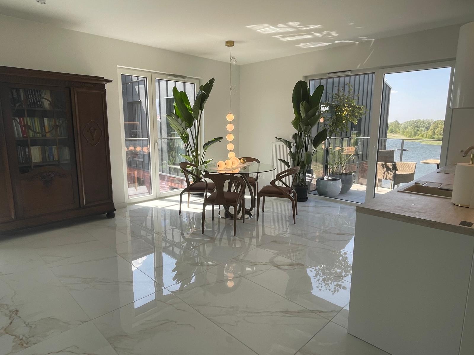 Jasne wnętrze z błyszczącymi płytkami podłogowymi, okrągłym stołem i roślinami w donicach przy oknach balkonowych z widokiem na jezioro. Klasyczna szafa z książkami.