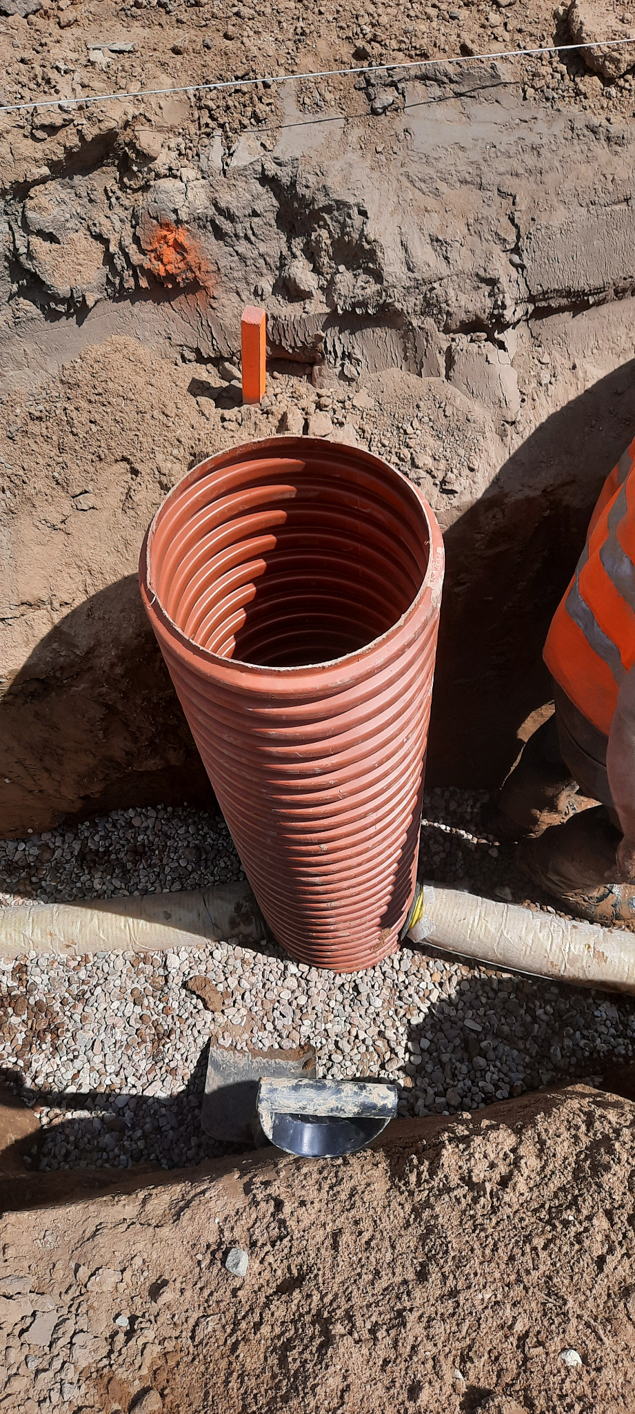 Pomarańczowa rura drenażowa w wykopie, podsypana kamieniami. Widoczny fragment rury oraz narzędzie. W tle ziemia i pomarańczowy słupek.