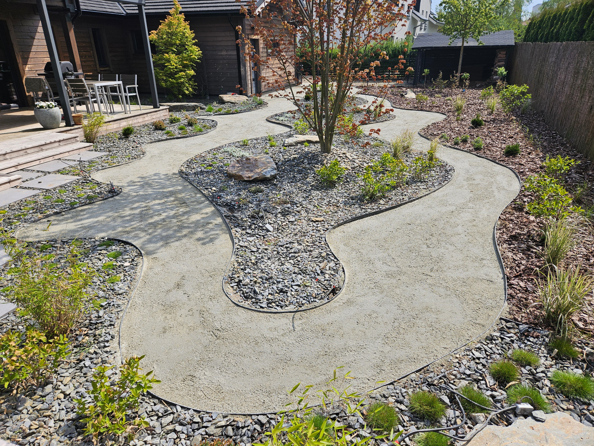 Kręta ścieżka z szarego kruszywa w ogrodzie, obrzeża z czarnego kamienia, wysypane korą rabaty z roślinami. W tle drewniana zabudowa.