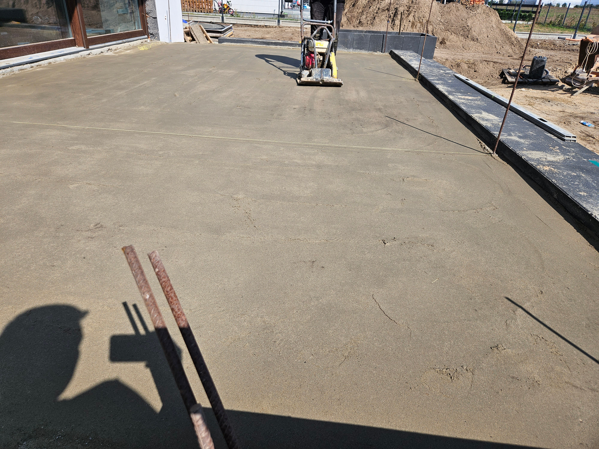 Ubijanie piasku zagęszczarką wibracyjną. Przygotowanie podłoża pod kostkę brukową. Widoczne szyny i równa powierzchnia piasku. Cień osoby fotografującej.