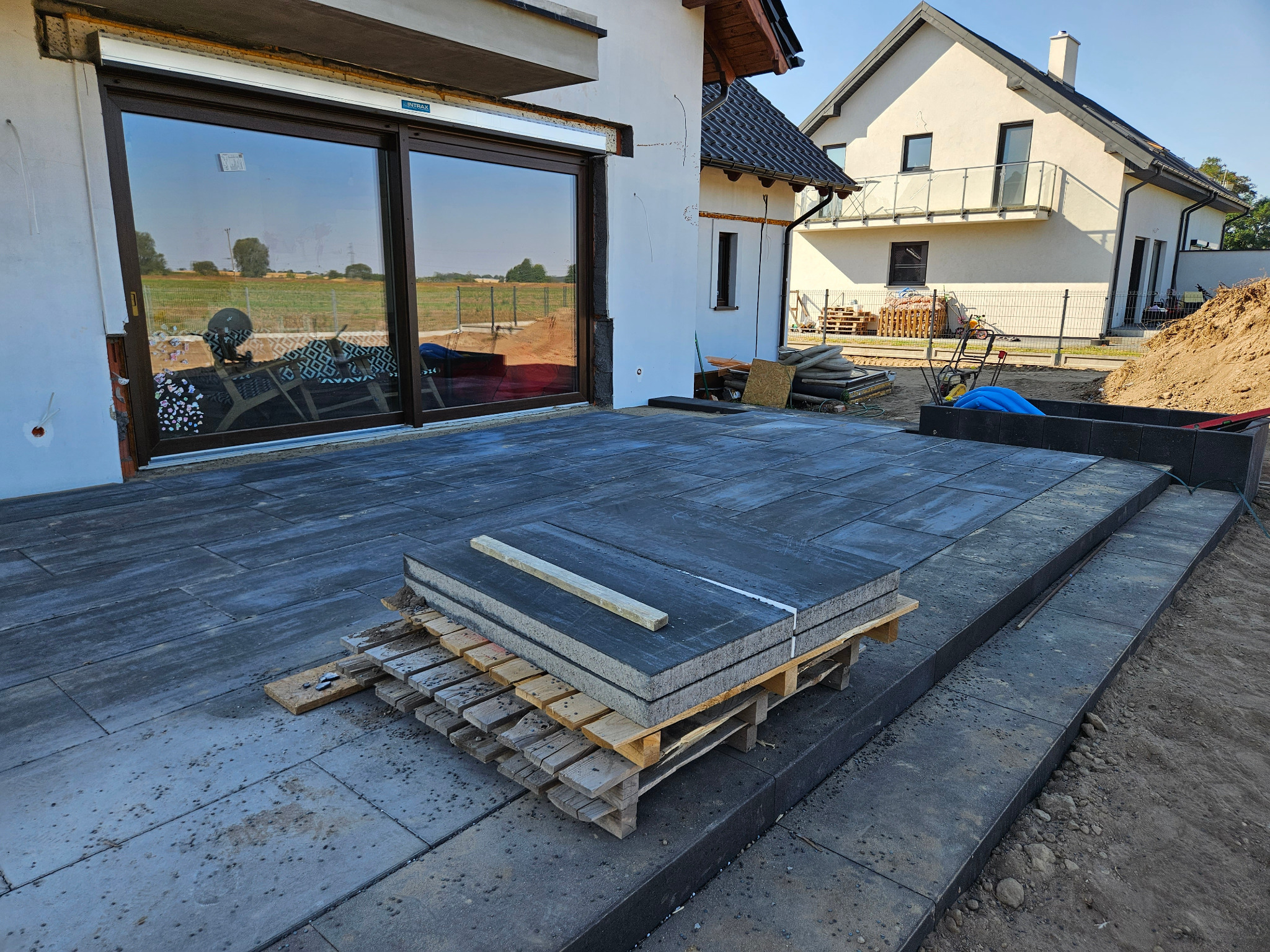 Ciemnoszara kostka brukowa ułożona przy budynku z dużym oknem, paleta z płytami na pierwszym planie. Widoczne przygotowania do dalszych prac.