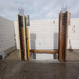 Matiitsiv - Zbrojenie pionowe w szalunku drewnianym, przygotowane do zalania betonem. Widoczne bloczki betonowe i fragment wylewki. Prace budowlane w toku, szare niebo w tle.