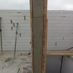 Wznoszenie żelbetowego słupa w szalunku drewnianym z widocznym zbrojeniem. Ściany z bloczków betonu komórkowego w tle. Element konstrukcyjny w trakcie budowy.