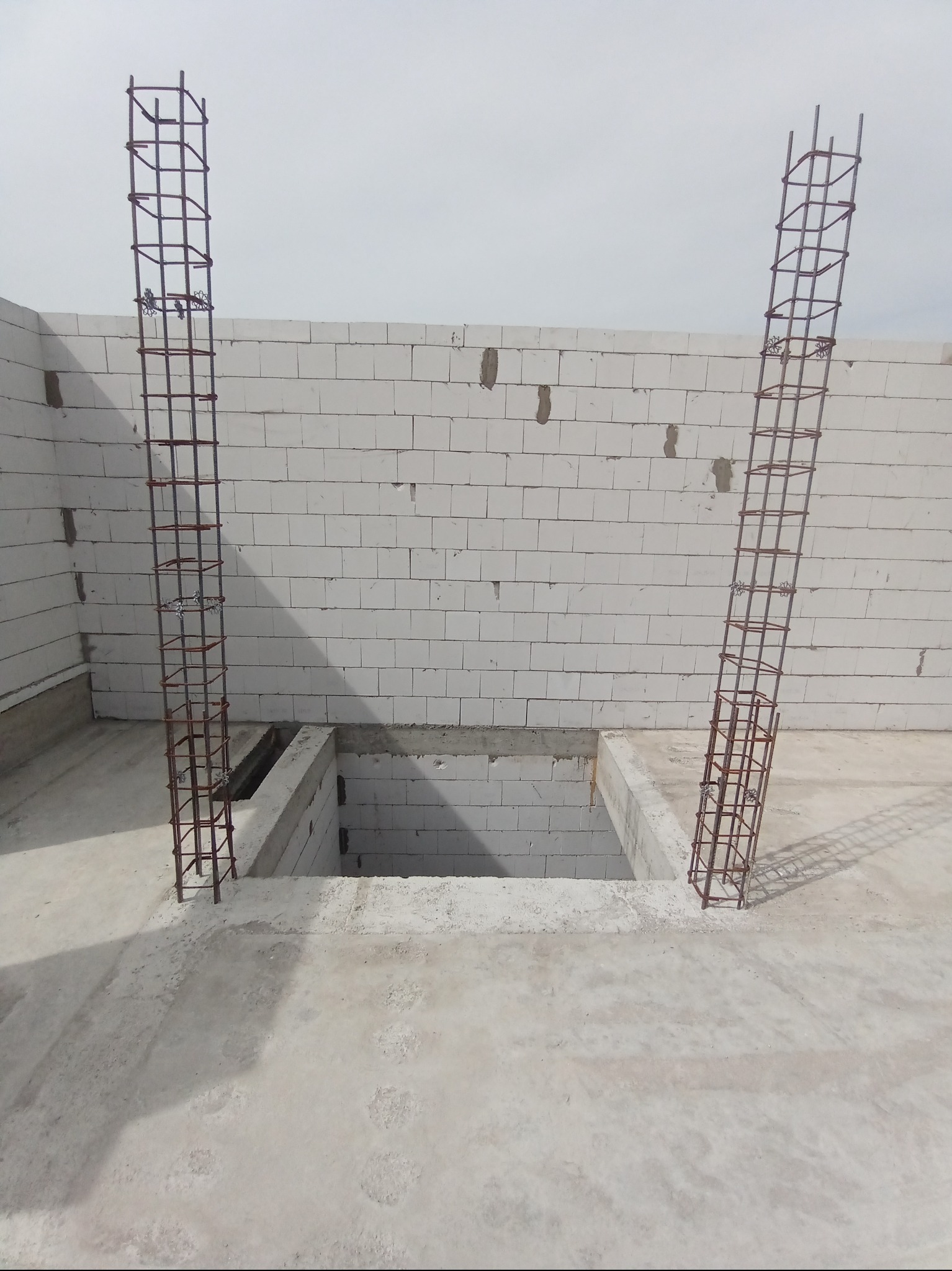 Dwie pionowe konstrukcje zbrojeniowe wystające z betonowej wylewki, ściany z białych bloczków, otwór w podłodze. Etap prac budowlanych.