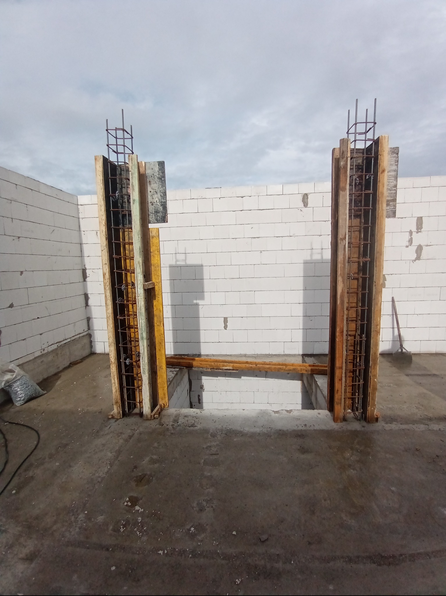 Zbrojenie pionowe w szalunku drewnianym, przygotowane do zalania betonem. Widoczne bloczki betonowe i fragment wylewki. Prace budowlane w toku, szare niebo w tle.