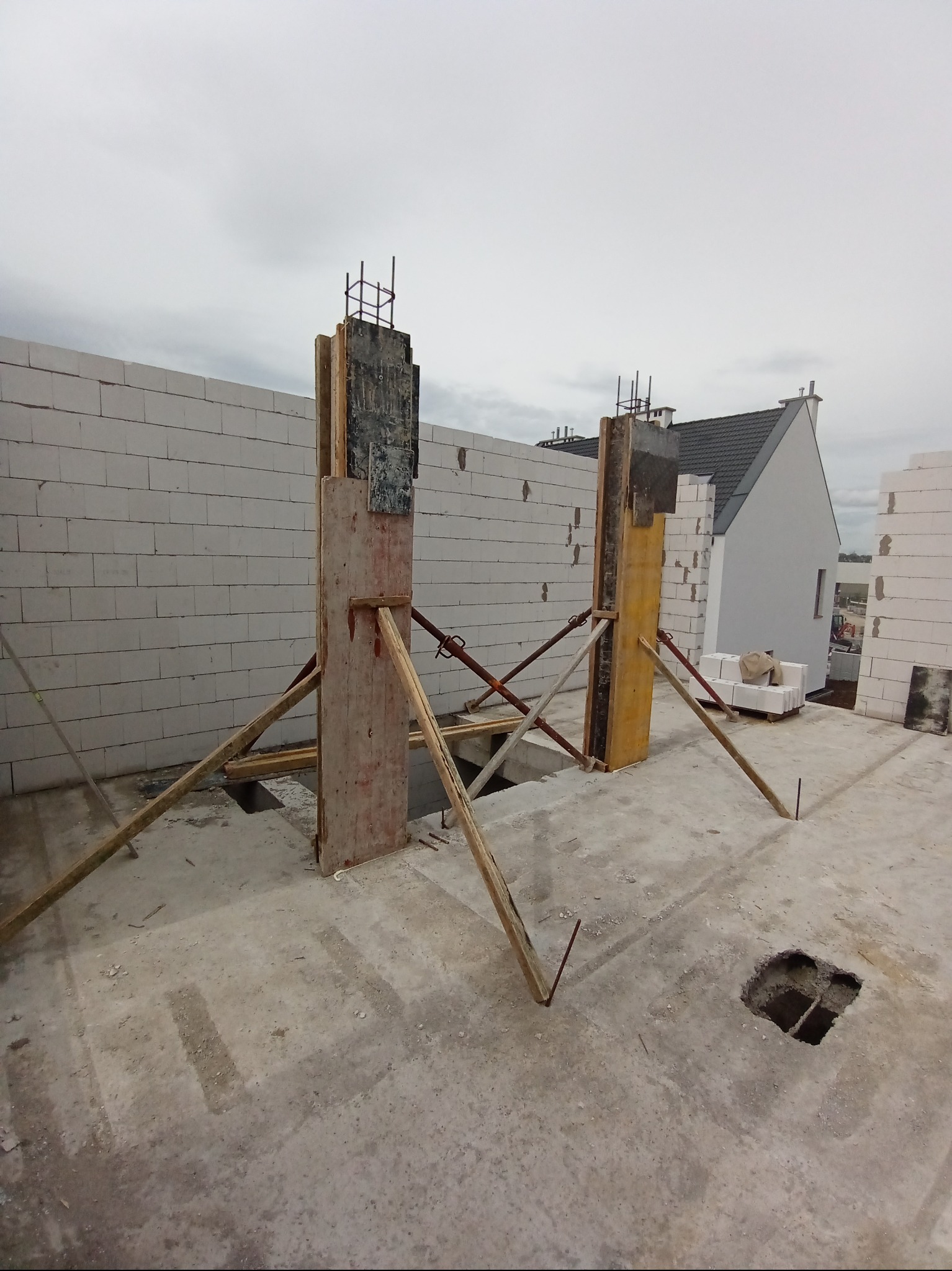 Szare, betonowe fundamenty z deskowaniem i zbrojeniem pionowym na budowie domu jednorodzinnego w pochmurny dzień. Widoczne bloczki betonowe i fragment domu w tle.