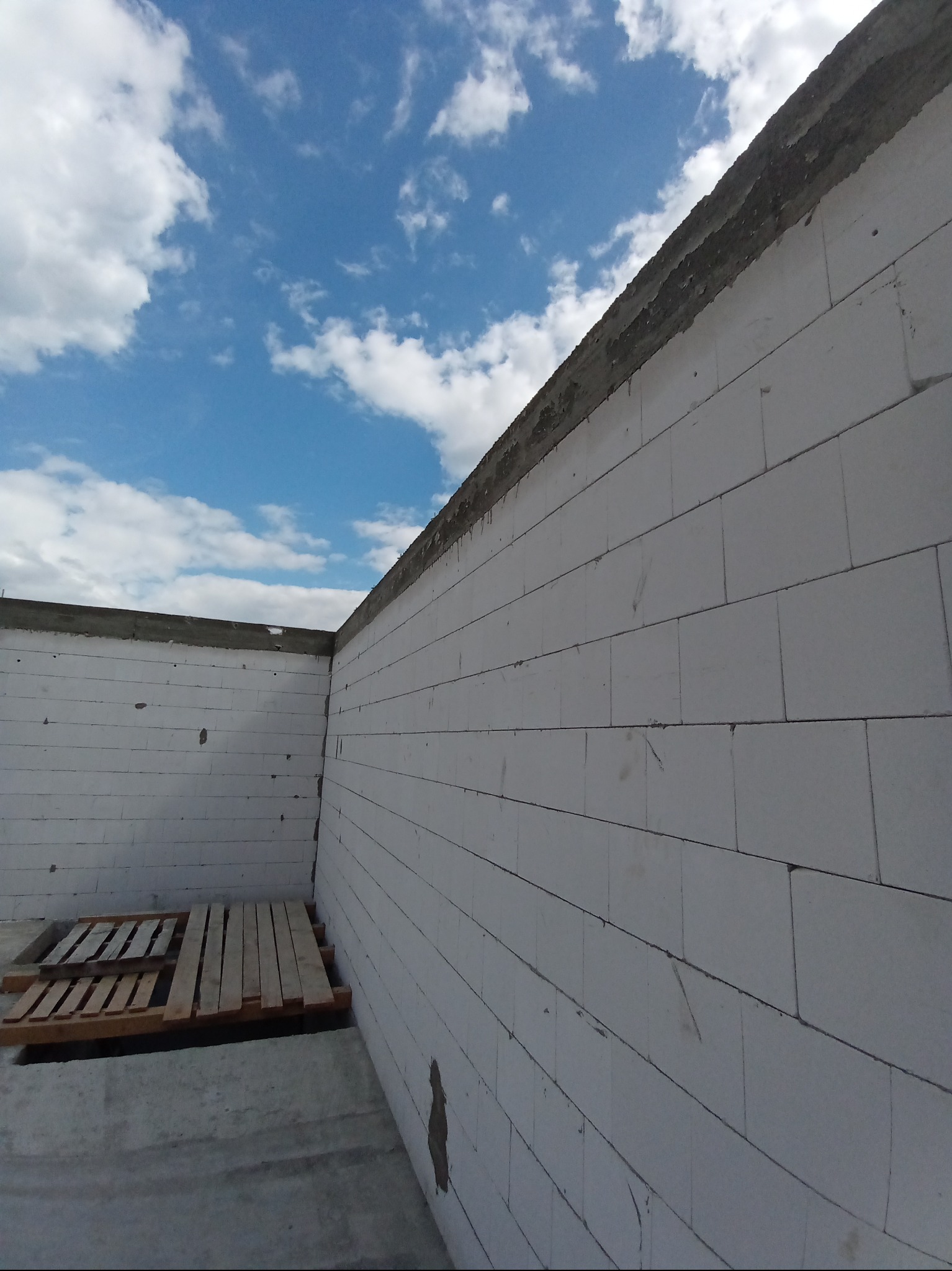 Wznoszenie ścian z bloczków betonu komórkowego na fundamencie. Widok z dołu na fragment muru i niebieskie niebo z chmurami. Paleta na posadzce.