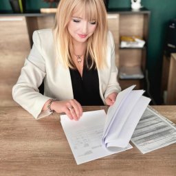 NOVELUM SPÓŁKA Z OGRANICZONĄ ODPOWIEDZIALNOŚCIĄ - Kobieta w biurze przegląda dokumenty na drewnianym biurku. Na ścianie zegar i dekoracje. Uśmiechnięta blondynka w białym żakiecie analizuje papiery.