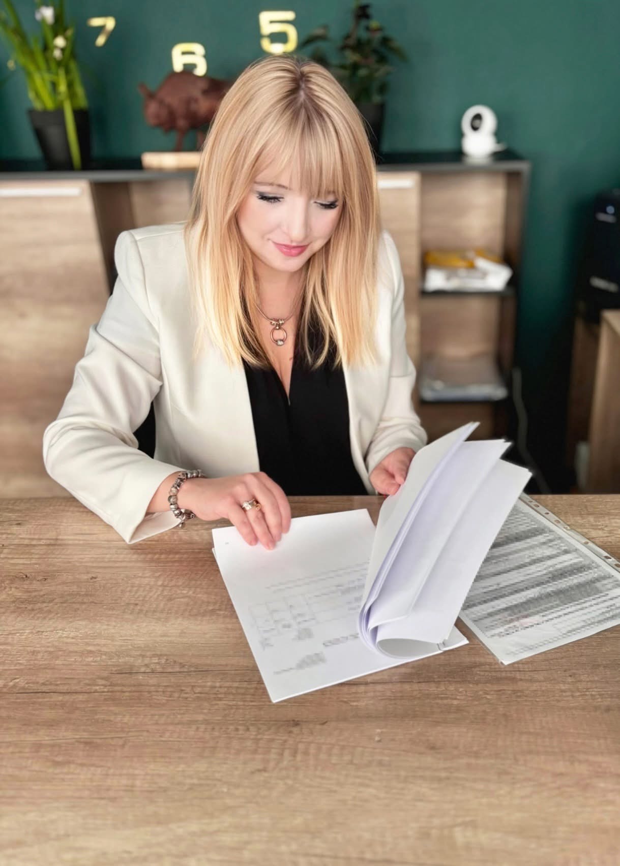 Kobieta w biurze przegląda dokumenty na drewnianym biurku. Na ścianie zegar i dekoracje. Uśmiechnięta blondynka w białym żakiecie analizuje papiery.