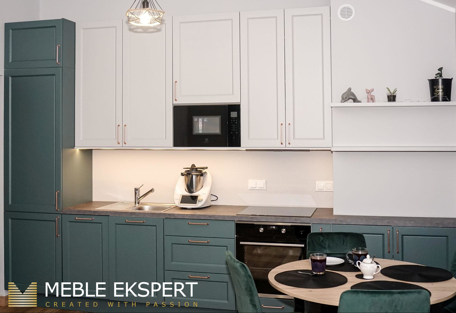 Nowoczesna kuchnia z zielonymi i białymi szafkami, blatem w odcieniach szarości i miedzianymi uchwytami. Widoczny stół z krzesłami i sprzęt AGD.