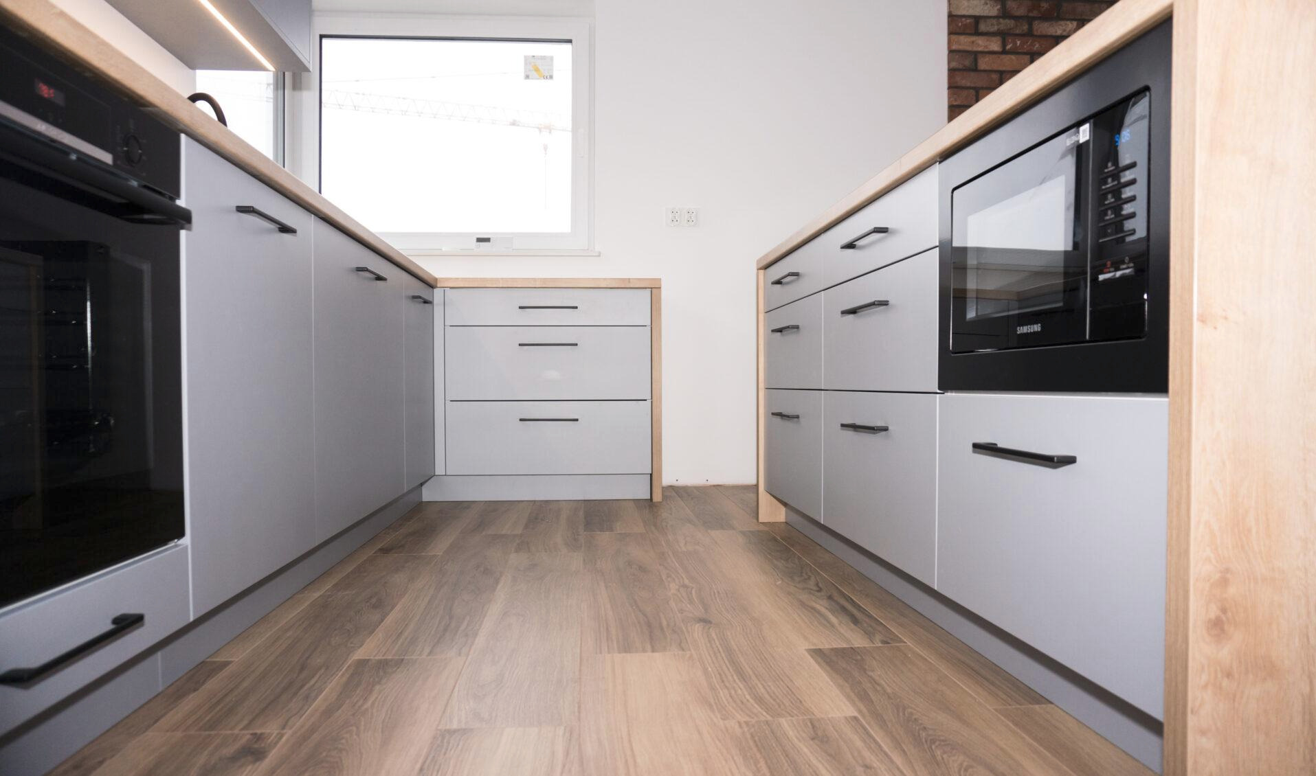 Nowoczesna kuchnia w szarościach z drewnianym blatem i czarnymi uchwytami, widok z poziomu podłogi, okno w tle, minimalistyczny design.