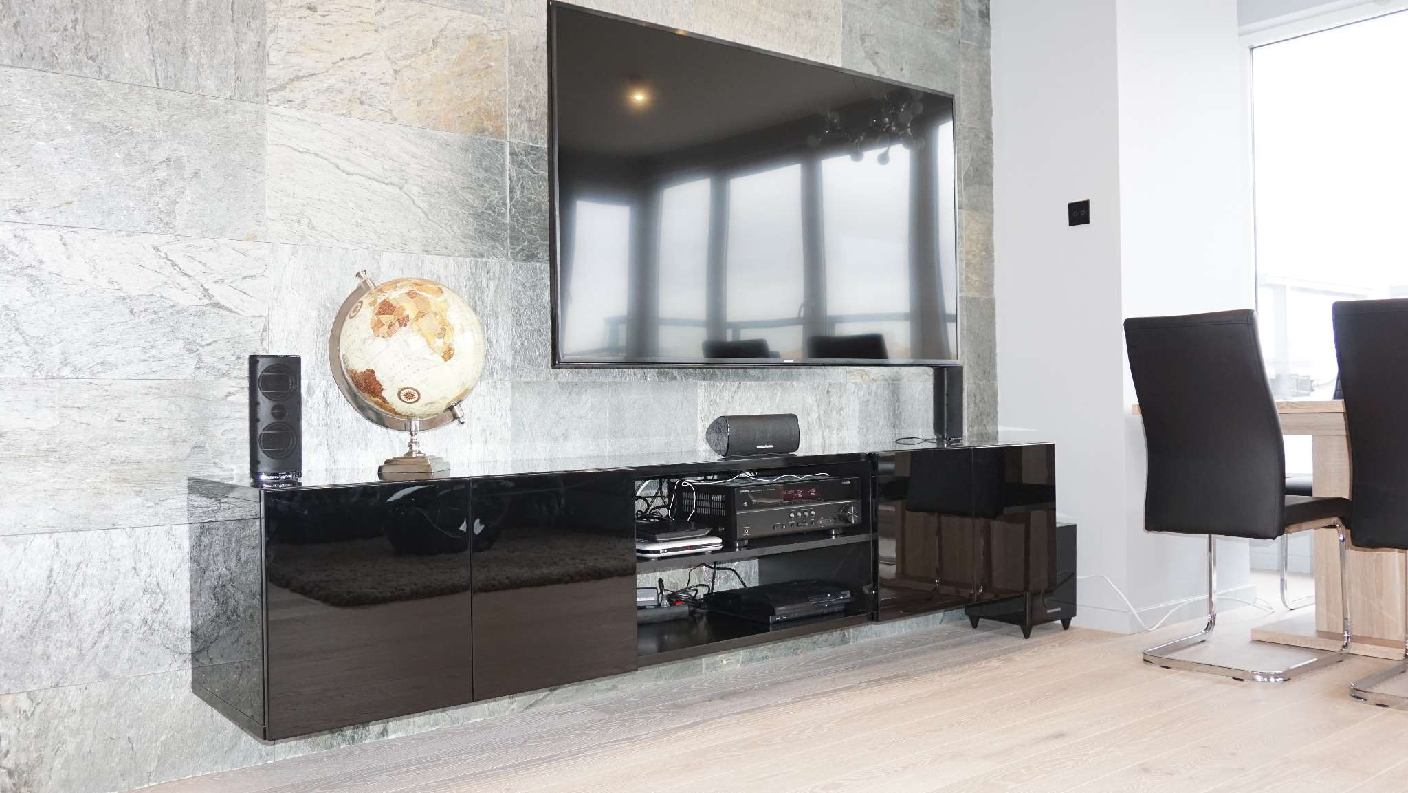 Czarna, lakierowana szafka RTV na tle ściany z kamienia, z globusem i głośnikiem. Nad nią zawieszony telewizor. W tle krzesła i okno.