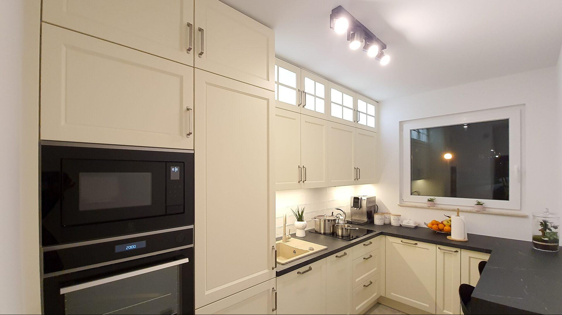 Jasna kuchnia z zabudową po sufit, czarnym blatem i sprzętem AGD w zabudowie. Nad blatem okno z widokiem na zewnątrz, oświetlenie punktowe na suficie.