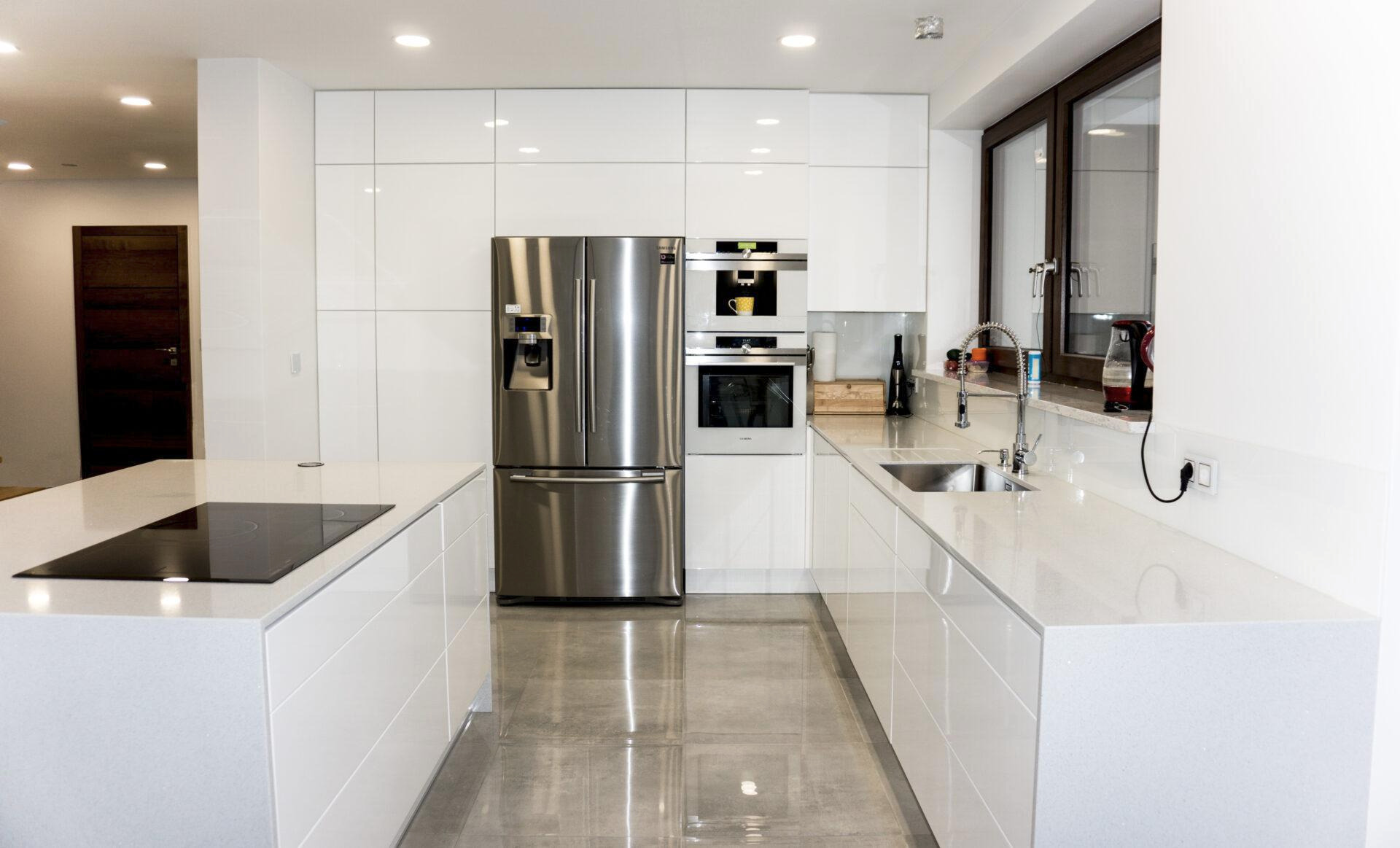 Nowoczesna, biała kuchnia z wyspą i sprzętem AGD ze stali nierdzewnej w Gdańsku. Minimalistyczny design, jasne wnętrze, widok na okno z ciemną ramą.