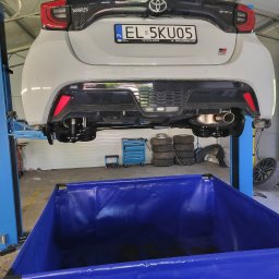 AutoRust Protect - Konserwacja podwozi - Biała Toyota Yaris GR Sport na podnośniku w warsztacie, widok z tyłu. Widoczny niebieski pojemnik na zużyte płyny, opony i narzędzia. Tablica rejestracyjna EL 5KU05.