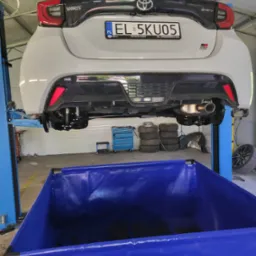 Biała Toyota Yaris GR Sport na podnośniku w warsztacie, widok z tyłu. Widoczny niebieski pojemnik na zużyte płyny, opony i narzędzia. Tablica rejestracyjna EL 5KU05.