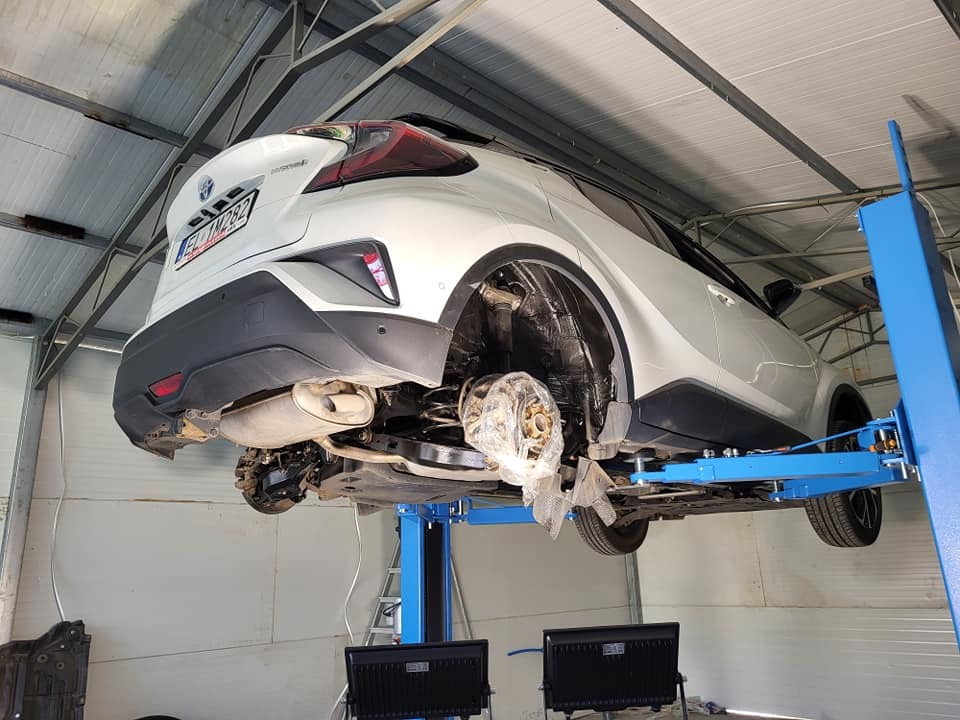 Biały samochód na podnośniku w warsztacie, widoczne zdjęte koła, blacha falista na suficie. C-HR Hybrid na niebieskim podnośniku.