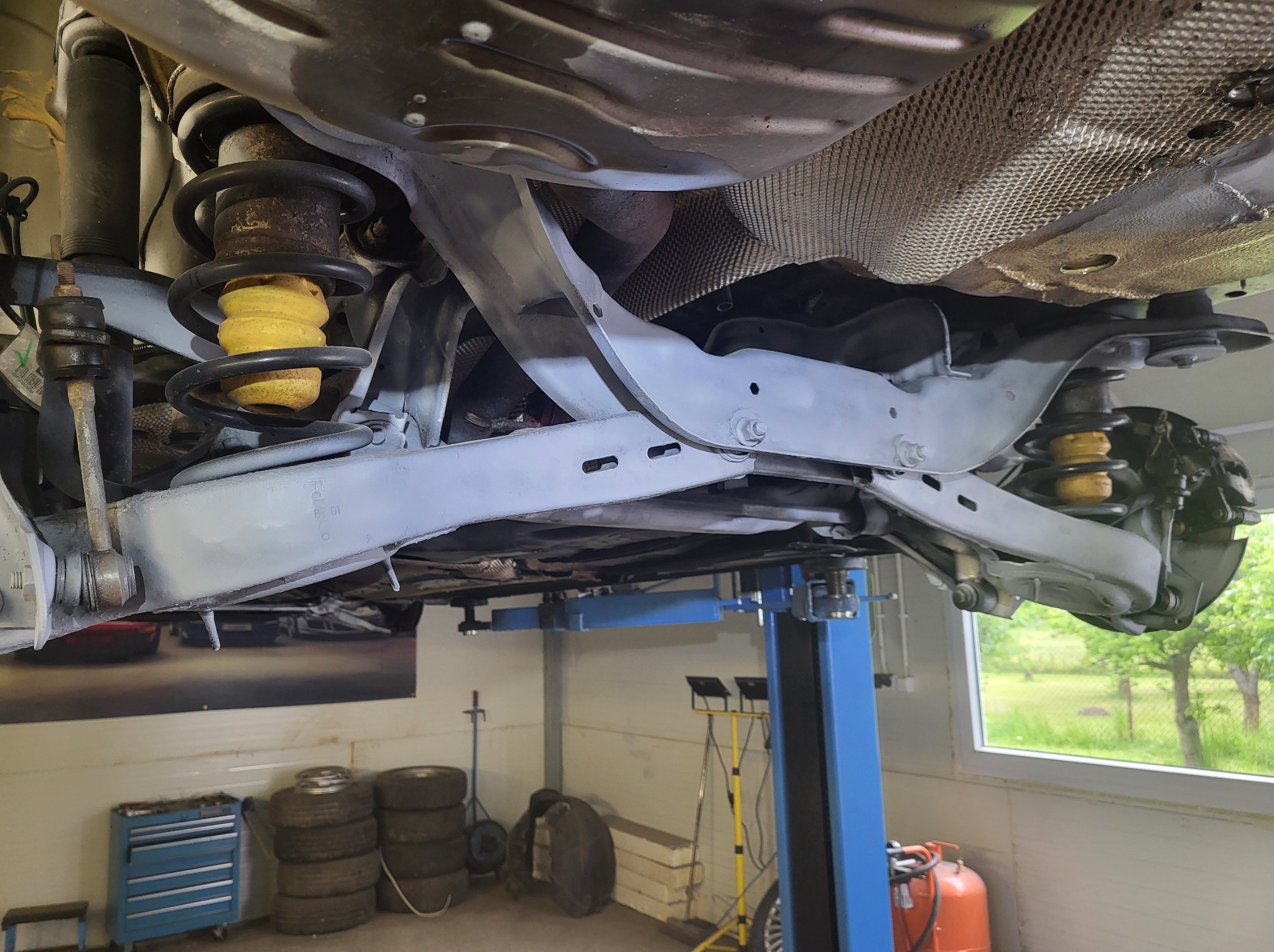 Podwozie auta po piaskowaniu i malowaniu, zawieszone na podnośniku w warsztacie. Widoczne elementy zawieszenia, sprężyny i amortyzatory. W tle narzędzia i opony.