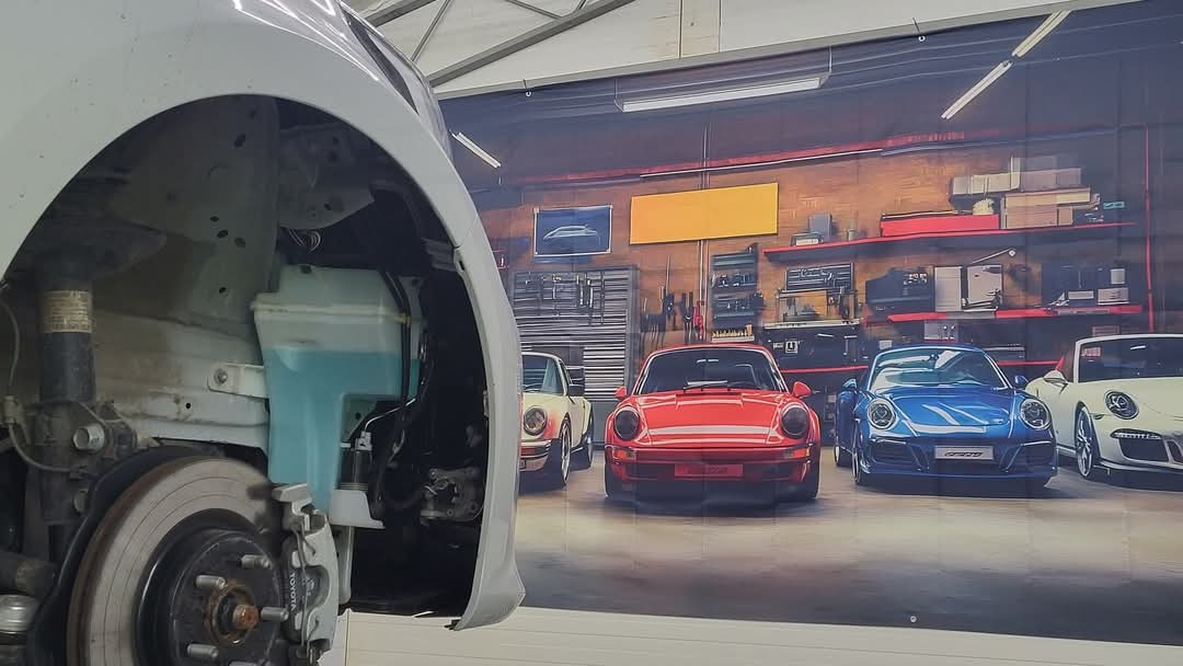 Rozłożony przód białego auta w warsztacie. Widoczne elementy zawieszenia i zbiornik płynu. W tle grafika z samochodami Porsche na tle warsztatu.
