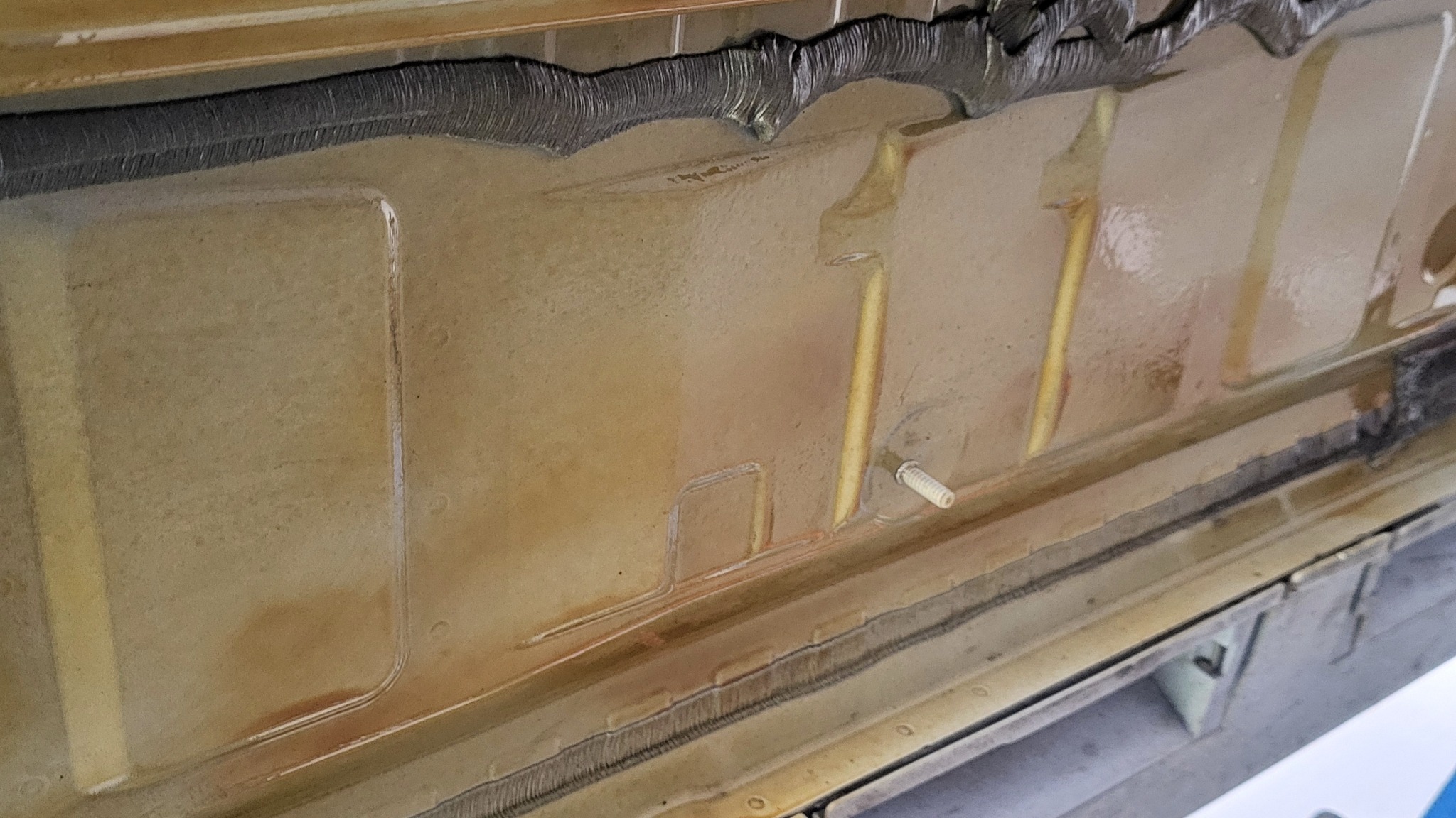 Wnętrze progu samochodu po zabezpieczeniu antykorozyjnym; widoczne ślady preparatu, wkręt mocujący i uszczelka. Ujęcie z bliska.