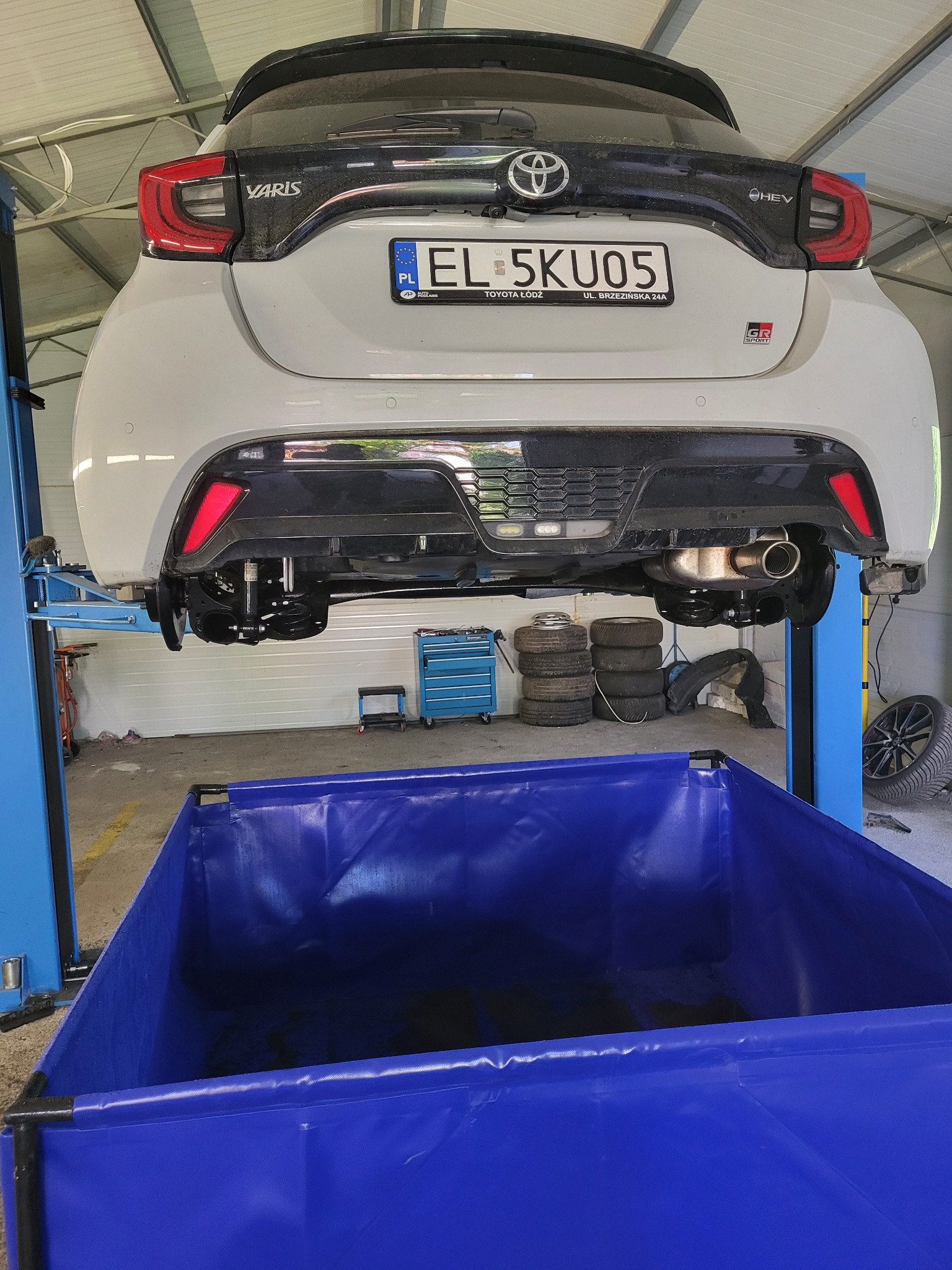 Biała Toyota Yaris GR Sport na podnośniku w warsztacie, widok z tyłu. Widoczny niebieski pojemnik na zużyte płyny, opony i narzędzia. Tablica rejestracyjna EL 5KU05.