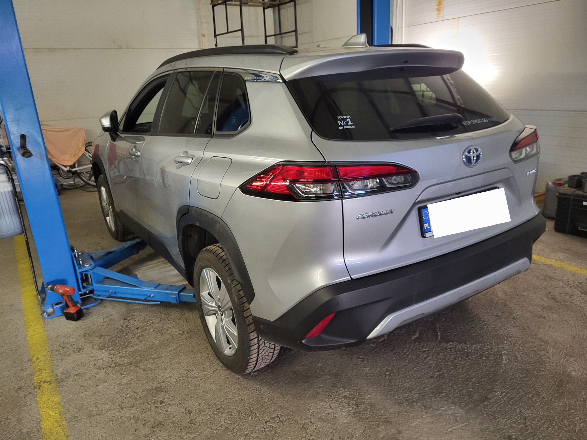 Srebrna Toyota Corolla Cross na podnośniku w warsztacie, widok z tyłu. Widoczny niebieski podnośnik i narzędzia. Na szybie naklejka 'Toyota Nr. 1'.
