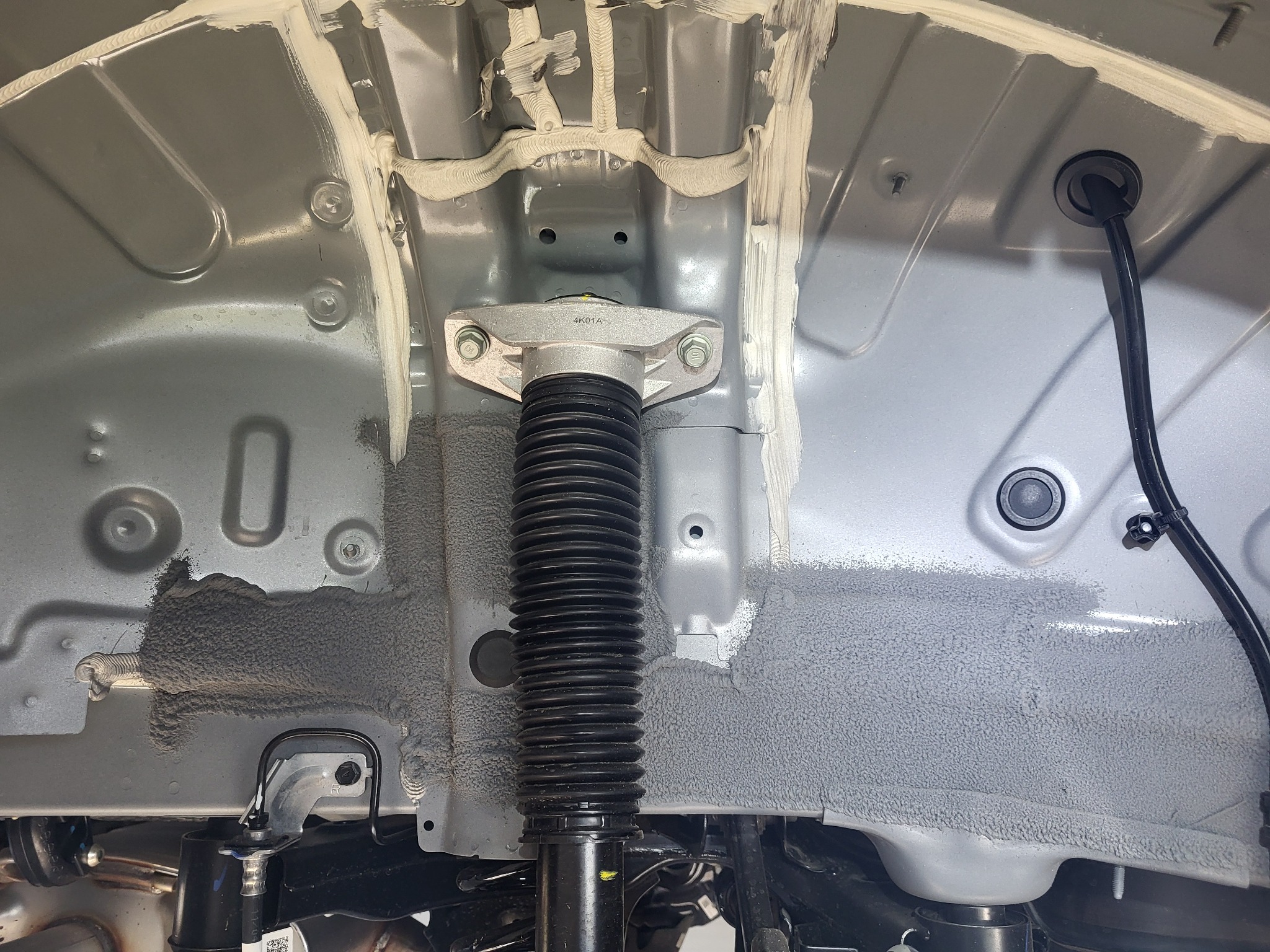 Wnętrze nadkola samochodu po nałożeniu warstwy ochronnej. Widoczny amortyzator, przewody oraz elementy karoserii zabezpieczone szarym preparatem antykorozyjnym.