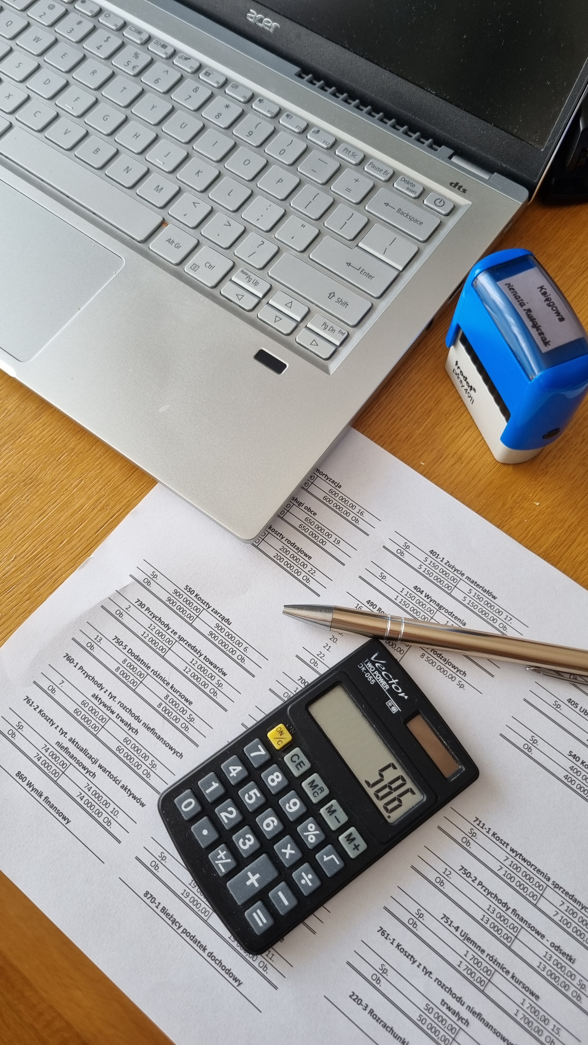 Kalkulator na dokumencie finansowym obok laptopa Acer i pieczątki z imieniem i nazwiskiem księgowej. Elementy biurowe na drewnianym biurku.