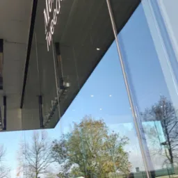 Refleksy drzew i auta w umytej szybie salonu Mercedes-Benz. Widoczny napis 'Mercedes Tweedenham' na górze okna, oraz fragment podnośnika do mycia okien.