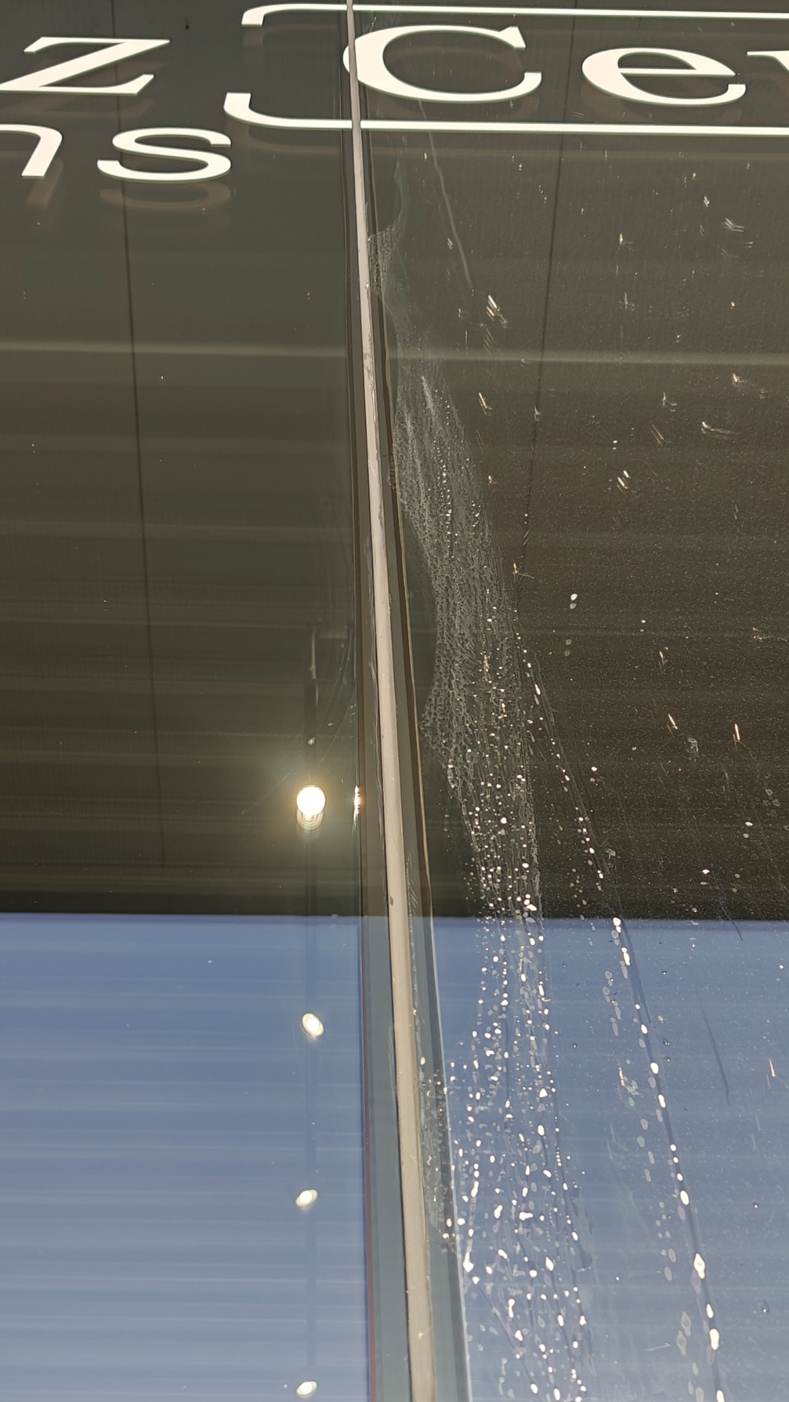 Brudna szyba okienna z zaciekami i plamami, widoczne odbicia światła i fragment napisu na elewacji. Szyba wymaga umycia, co poprawi jej wygląd i przejrzystość.