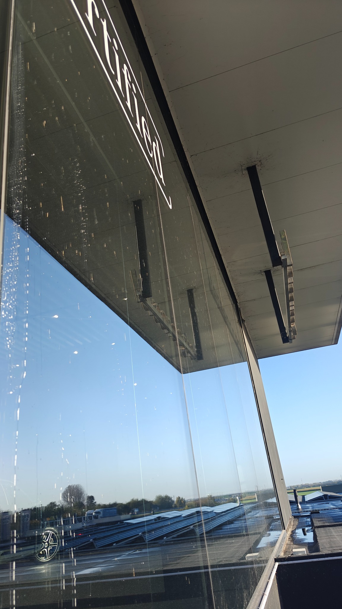 Szklana fasada budynku z widocznymi smugami po myciu, odbijająca błękitne niebo i otoczenie. Perspektywa z dołu ukazuje konstrukcję dachu i czystość szyb.