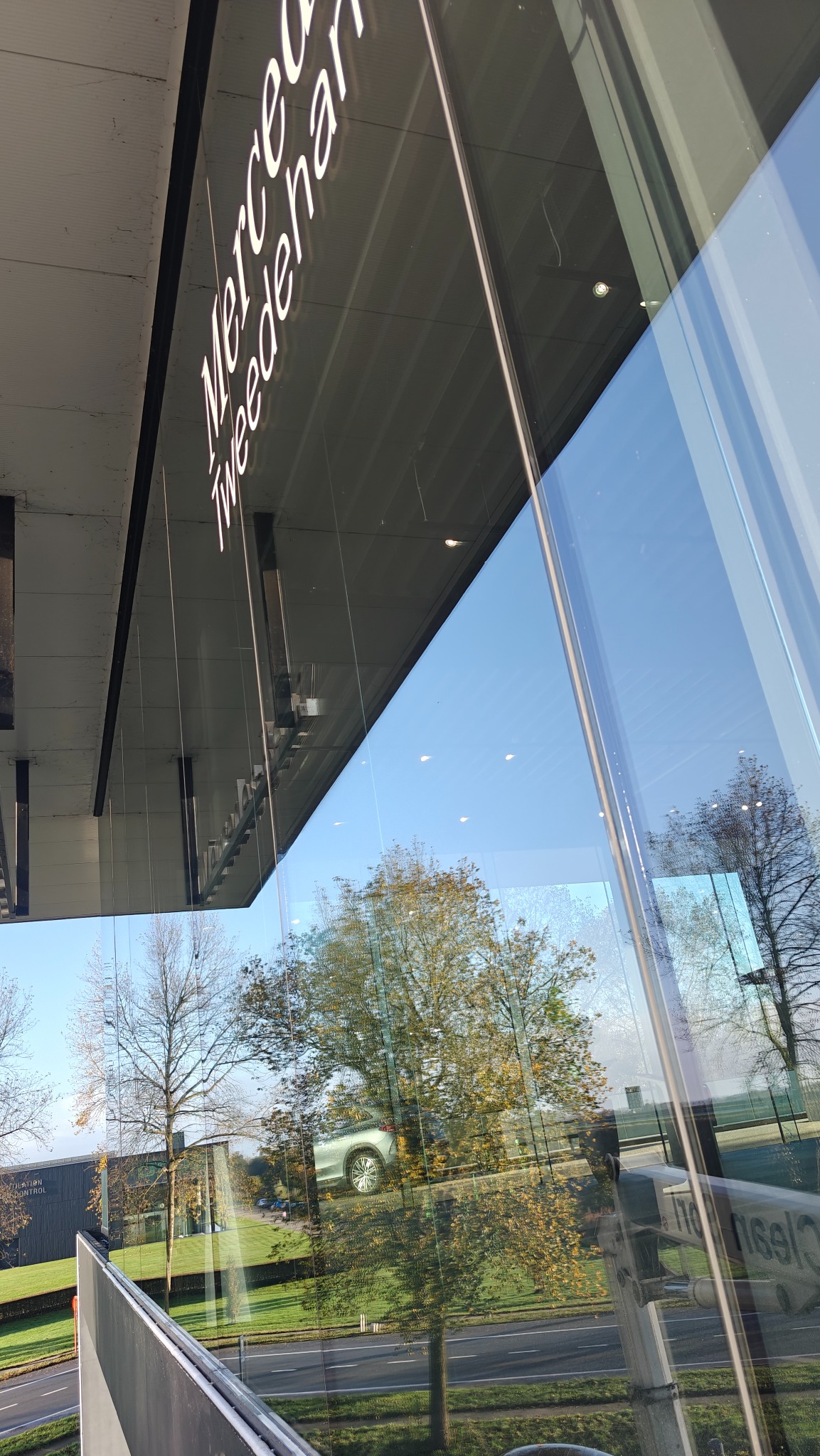 Refleksy drzew i auta w umytej szybie salonu Mercedes-Benz. Widoczny napis 'Mercedes Tweedenham' na górze okna, oraz fragment podnośnika do mycia okien.