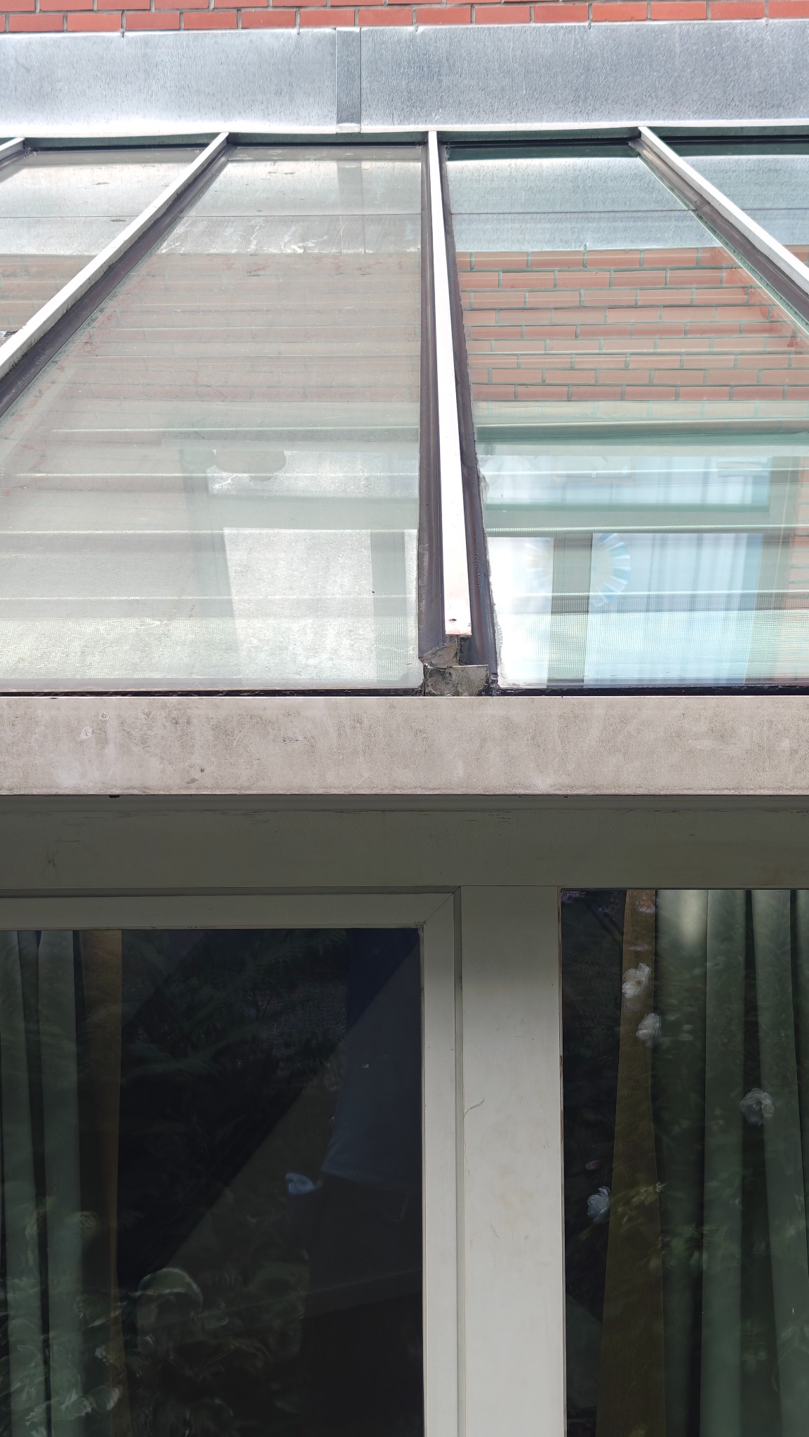 Zabrudzona, przeszklona konstrukcja dachu z widocznymi smugami i osadami. W tle ceglany budynek i okna z zasłonami. Widoczne zacieki i niedoskonałości.