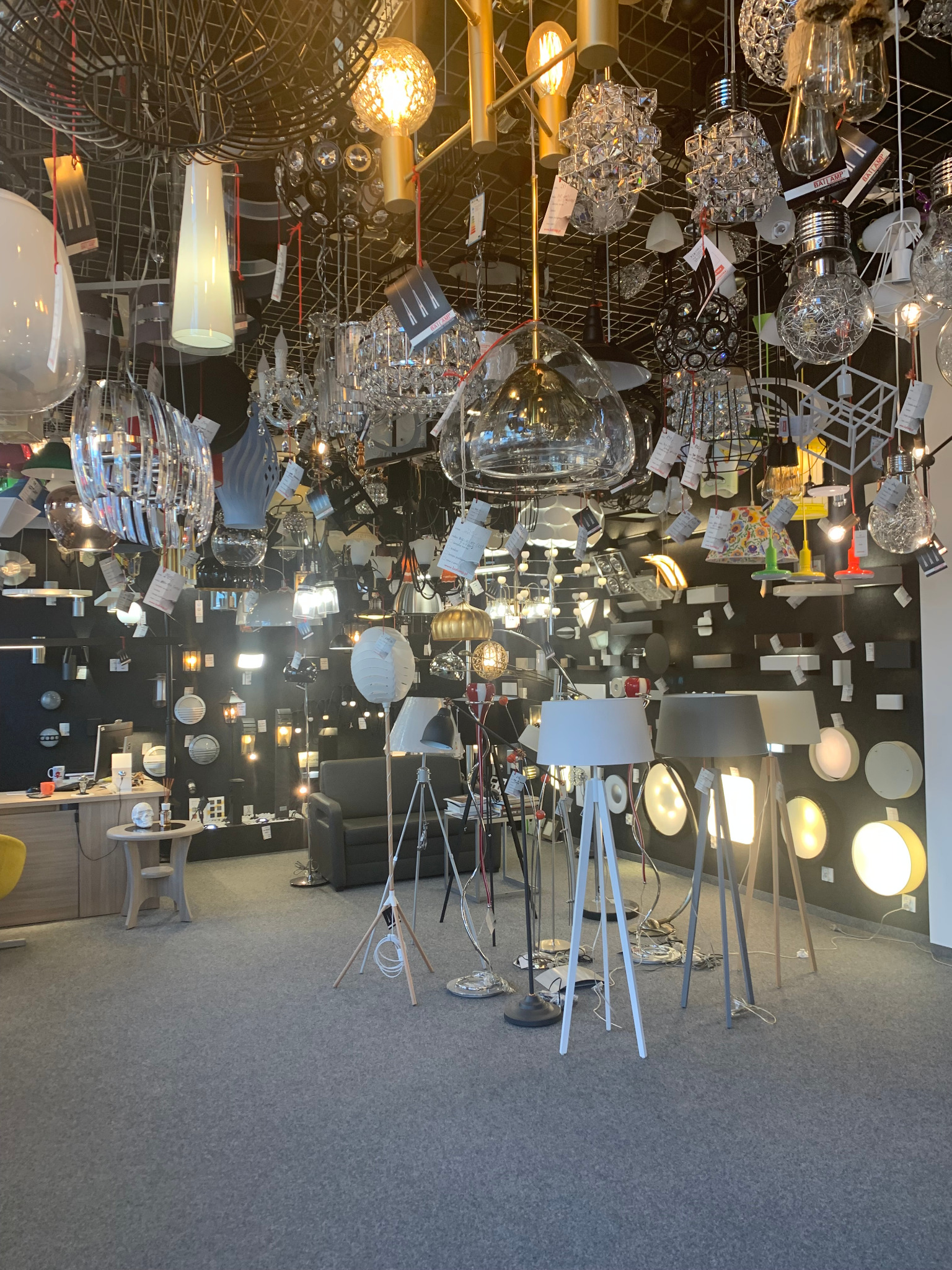 Wnętrze sklepu z oświetleniem: różnorodne lampy wiszące i stojące, ekspozycja na ścianie, szara podłoga. Duży wybór lamp w jednym miejscu.