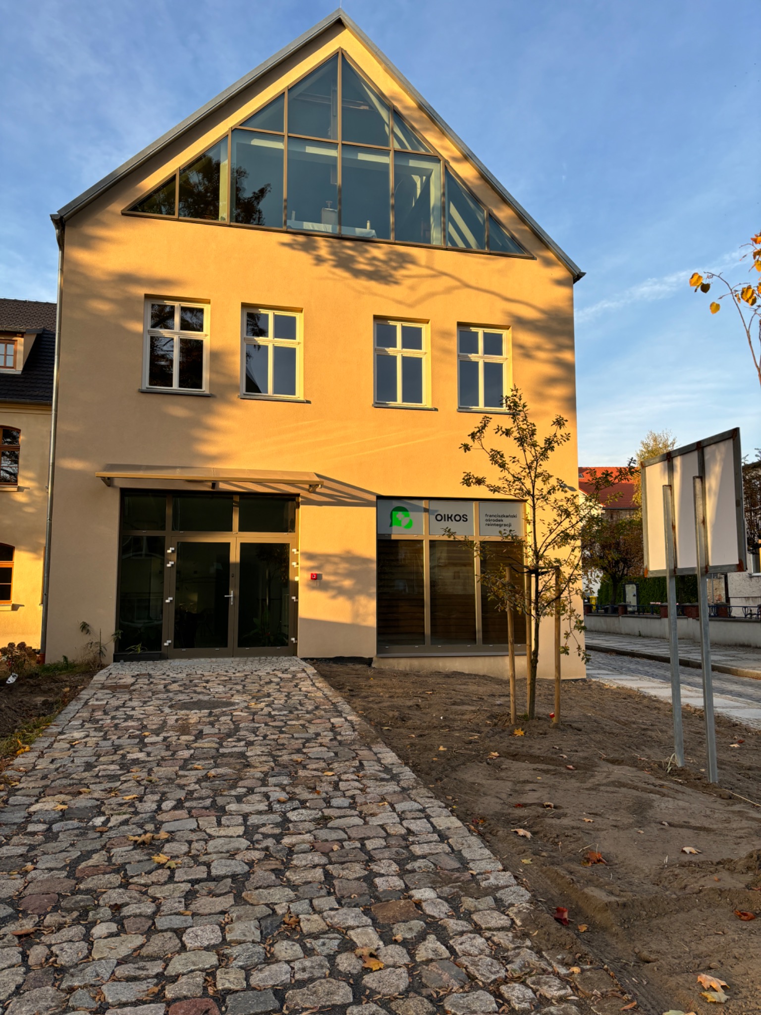 Budynek Oikos, Franciszkański Ośrodek Reintegracji, z charakterystycznym trójkątnym oknem na poddaszu. Brukowany chodnik prowadzi do wejścia.