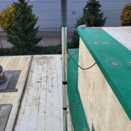 Net-Tech Marcin Kasprzak - Antena Starlink zamontowana na dachu budynku, widok z dołu. Szara elewacja w tle, zielone obramowanie dachu. Kabel antenowy poprowadzony wzdłuż rury mocującej.