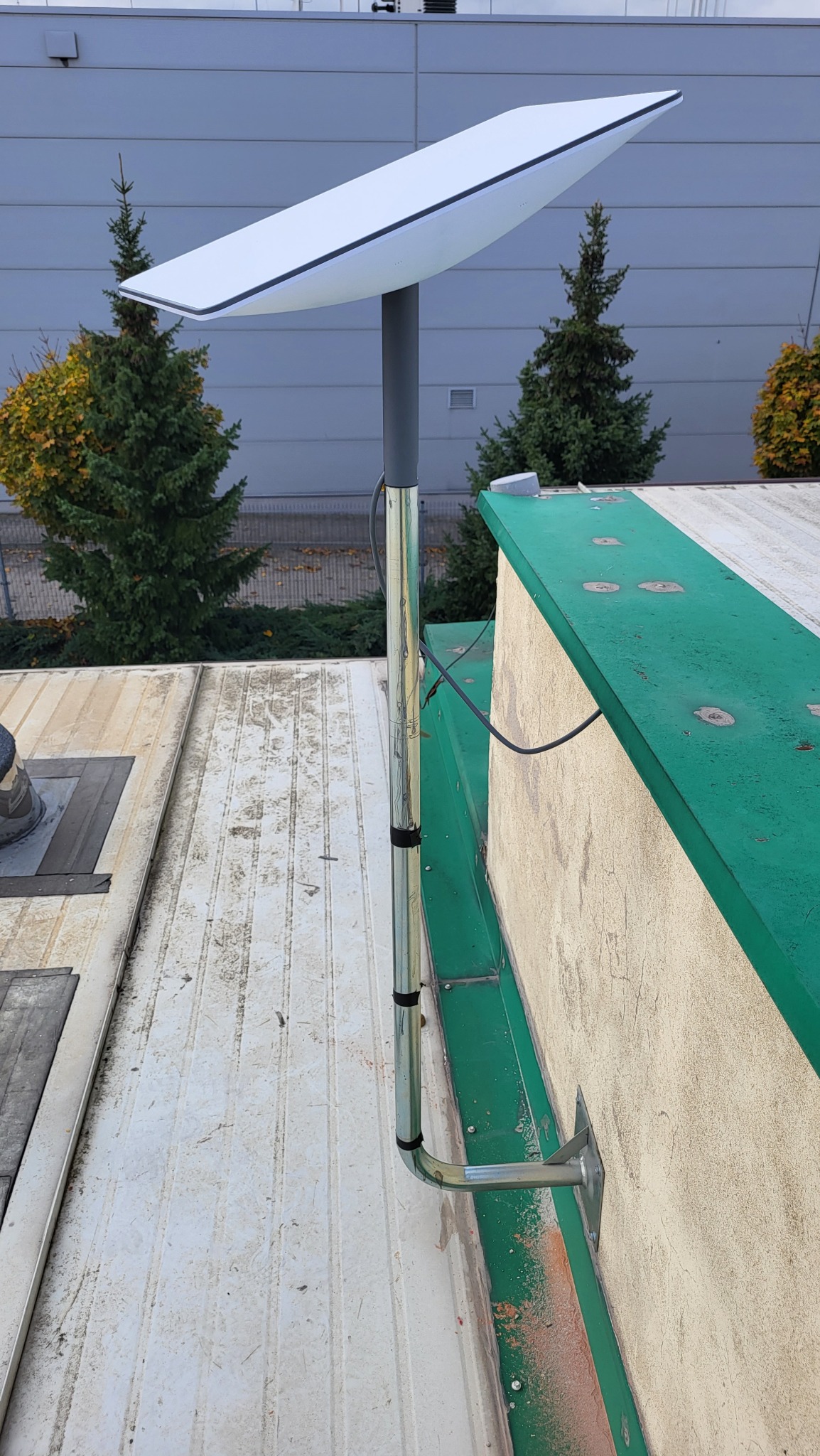 Antena Starlink zamontowana na dachu budynku, widok z dołu. Szara elewacja w tle, zielone obramowanie dachu. Kabel antenowy poprowadzony wzdłuż rury mocującej.
