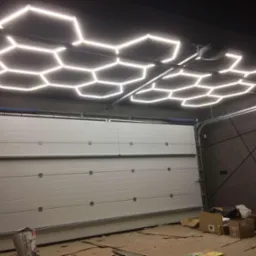 Oświetlenie LED w kształcie plastra miodu na suficie garażu z białą bramą segmentową. Widoczne kartony i worek z zaprawą. Nietypowy design oświetlenia.