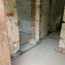 Surowy stan wnętrza z instalacją elektryczną: kable w korytach podłogowych, puszki w murze. W tle widoczne surowe ściany z cegły i betonu komórkowego.