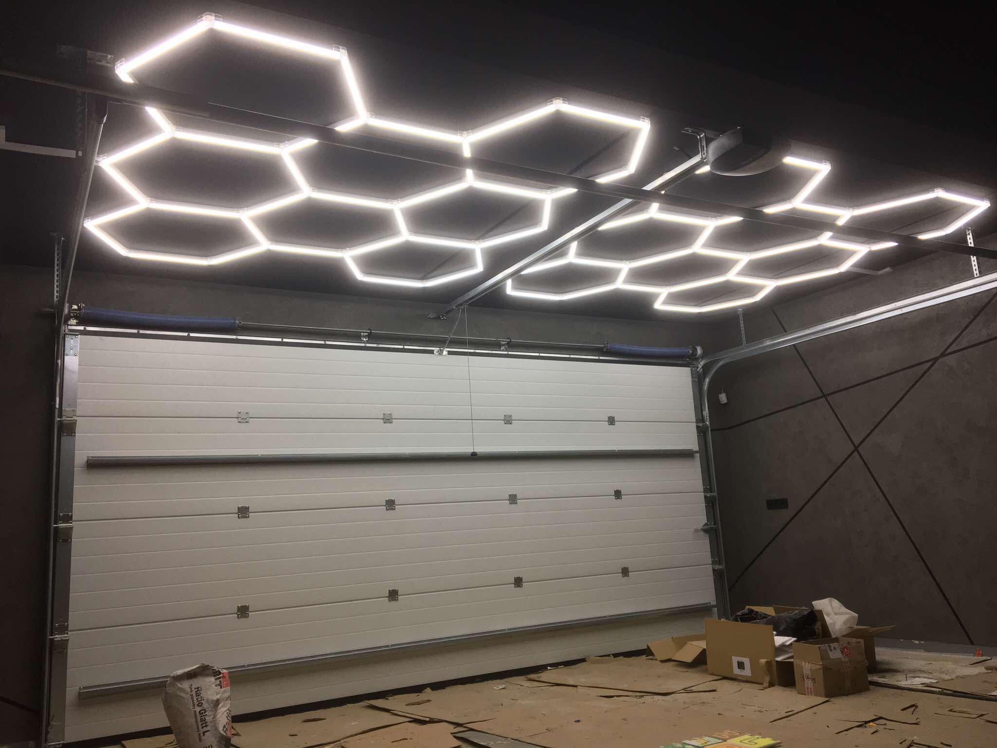 Oświetlenie LED w kształcie plastra miodu na suficie garażu z białą bramą segmentową. Widoczne kartony i worek z zaprawą. Nietypowy design oświetlenia.